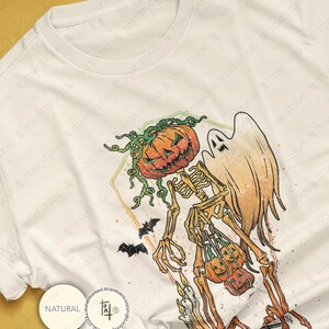 May include: Off-white t-shirt featuring a Halloween-themed graphic. The design includes a skeleton with a pumpkin head, a ghost, bats, and pumpkins. The graphic is in orange, green, and black. The shirt has a round neckline.