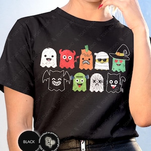 May include: Black t-shirt featuring a colorful Halloween-themed graphic. The design includes various cartoon ghosts and monsters in different colors and expressions. The shirt has a round neckline and short sleeves. The word "BLACK" is printed on a black circle.