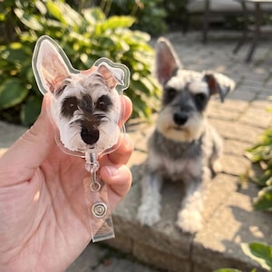 May include: A clear acrylic badge reel featuring a printed portrait of a Schnauzer dog with grey, black, and white fur. The circular-shaped topper displays the dog's face with pointed ears, while a retractable cord connects to a clear plastic strap.