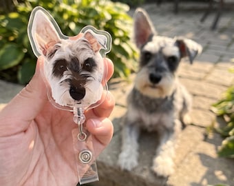 May include: A clear acrylic badge reel featuring a printed portrait of a Schnauzer dog with grey, black, and white fur. The circular-shaped topper displays the dog's face with pointed ears, while a retractable cord connects to a clear plastic strap.