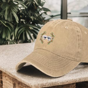 May include: A tan baseball cap with an embroidered design of two dogs wearing a wreath of green leaves.