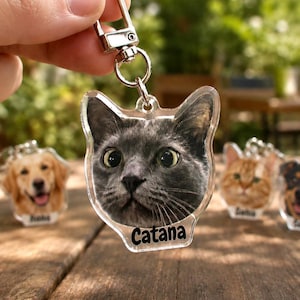 Puede incluir: Un llavero de acrílico transparente con una impresión fotográfica de la cara de un gato gris y el nombre Catana impreso en negro en la parte inferior. El llavero incluye un cierre de mosquetón de metal plateado y una argolla.