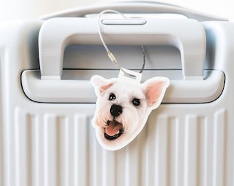 May include: A white luggage tag shaped like a dog's head, featuring a smiling expression with its tongue out. The tag is attached to a white suitcase handle with a silver wire. The dog has pink ears and a black nose.