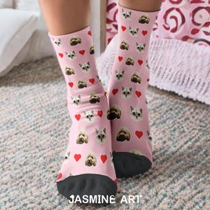 May include: Pink socks with a pattern of cat and dog faces and red hearts. The socks have black toes and the text "JASMINE ART" at the bottom. The socks are on a patterned rug.