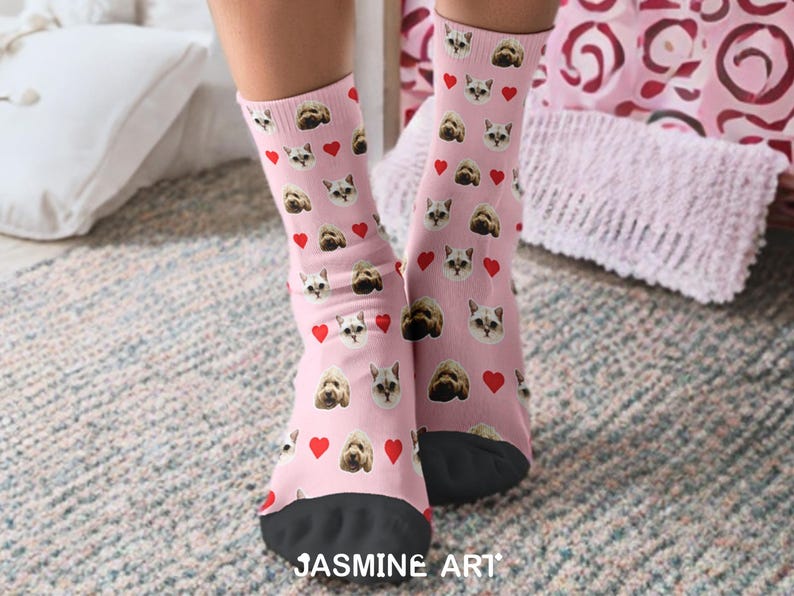 May include: Pink socks with a pattern of cat and dog faces and red hearts. The socks have black toes and the text "JASMINE ART" at the bottom. The socks are on a patterned rug.