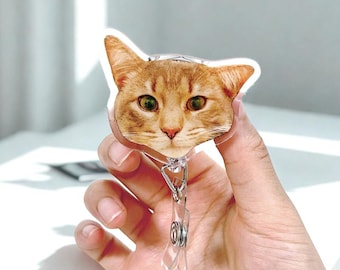 May include: A retractable badge holder featuring a realistic orange tabby cat face. The cat has green eyes and a pink nose. The badge reel has a clear plastic strap and a metal clip. The cat image is printed on a clear plastic badge.