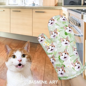Peut inclure: Un gant de cuisine vert avec un motif floral représentant un visage de chat répété sur toute la surface. Le chat a du poil blanc et des marques brunes. Le gant est dans un décor de cuisine.