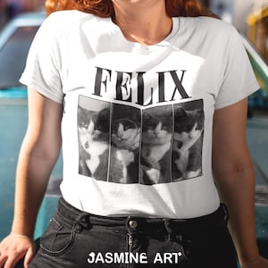 May include: White t-shirt featuring a black and white image of a cat with the word "FELIX" printed in black letters above the image. The text "JASMINE ART" is printed in black letters at the bottom of the shirt.