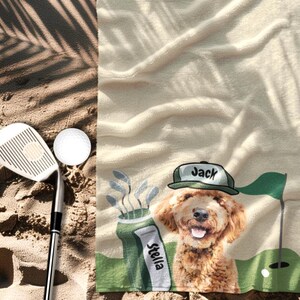 May include: A beige golf towel with a cartoon dog wearing a green hat that says "Jack". The towel has a golf course scene with a golf ball, flag, and golf club. A golf club and ball are on the sand.