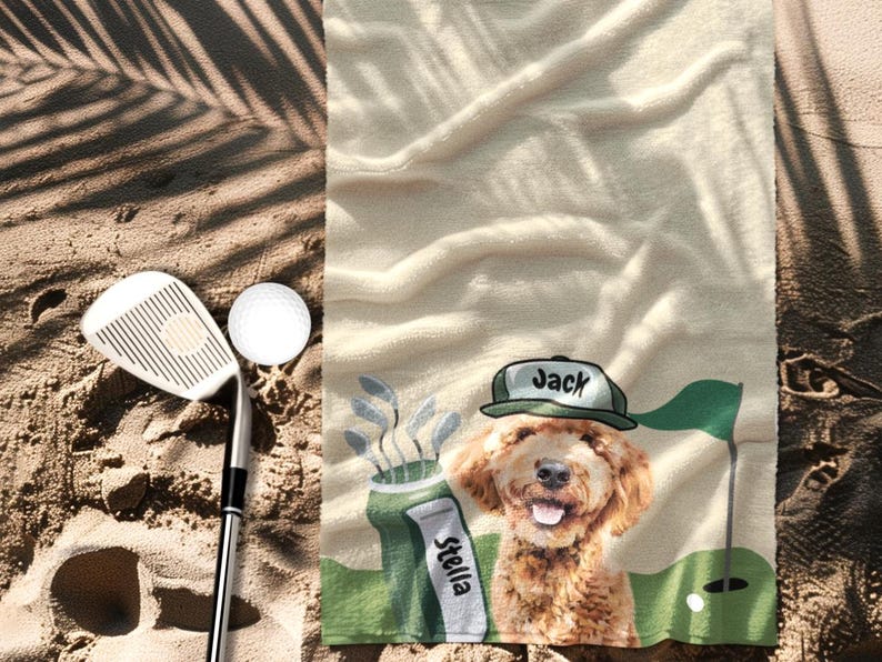 May include: A beige golf towel with a cartoon dog wearing a green hat that says "Jack". The towel has a golf course scene with a golf ball, flag, and golf club. A golf club and ball are on the sand.