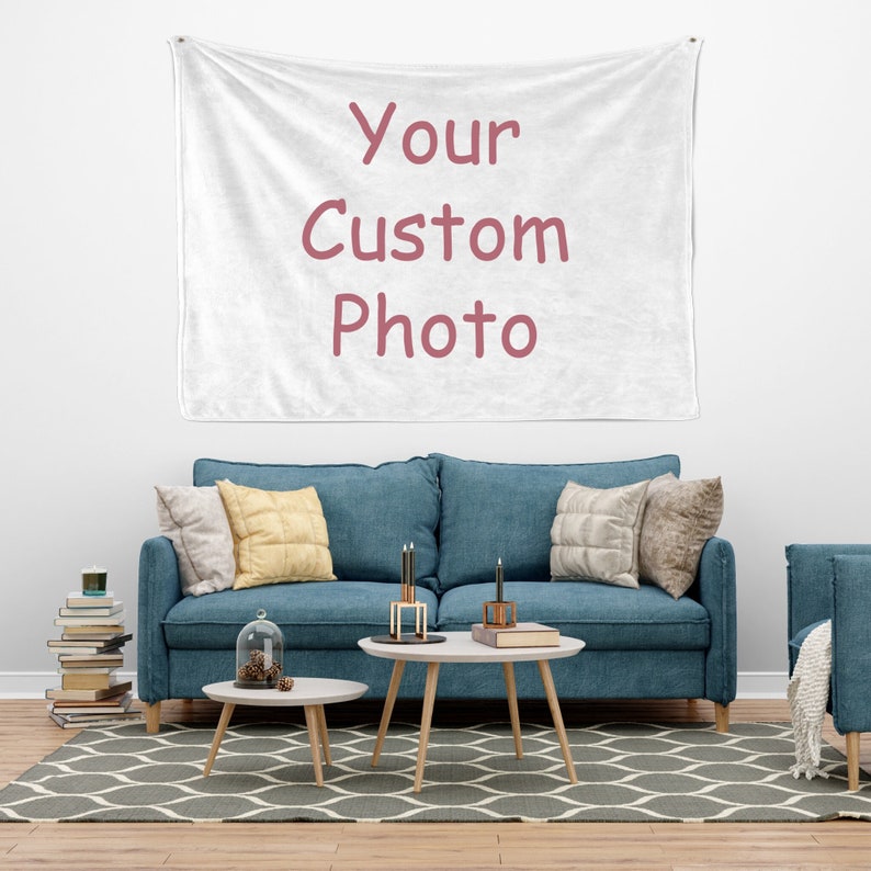 May include: A white throw blanket with the text "Your Custom Photo" in pink lettering hangs on a white wall above a blue sofa with pillows. The sofa is in a living room setting with a coffee table and side table.