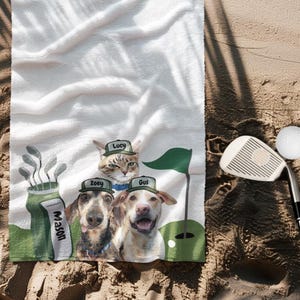 May include: White golf towel featuring a whimsical design with a cat and two dogs wearing golf hats, with the names "Lucy", "Zoey", and "Gus" printed on the hats. A golf club and ball are also visible.