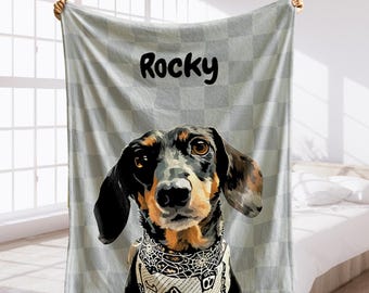 May include: A personalized blanket featuring a dachshund dog portrait with the name "Rocky" in black lettering. The dog is wearing a bandana with a spiderweb design. The background is a light gray checkered pattern.
