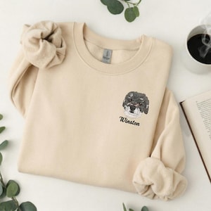 Peut inclure: Un sweat-shirt beige avec un col rond et des manches longues. Le sweat-shirt présente un portrait brodé d'un chien avec une fourrure noire et grise, et le nom "Winston" brodé sous l'image du chien. Le sweat-shirt est plié.