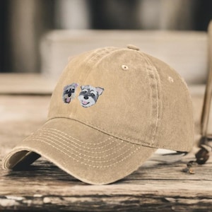 May include: A tan baseball cap with a distressed, washed-out finish. The front of the cap features a small, embroidered graphic of two Schnauzer dog faces, each with grey and white fur, black noses, and open mouths showing their tongues.