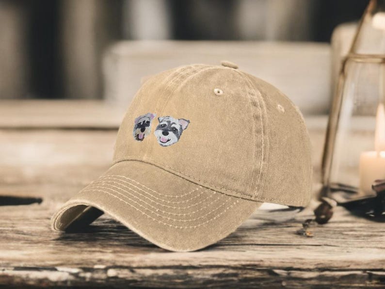 May include: A tan baseball cap with a distressed, washed-out finish. The front of the cap features a small, embroidered graphic of two Schnauzer dog faces, each with grey and white fur, black noses, and open mouths showing their tongues.