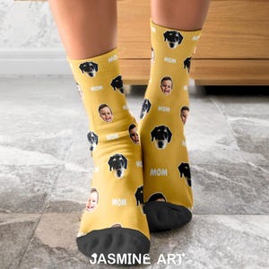 Op de afbeelding: Mosterdgele sokken met een herhalend patroon van een hondengezicht, een babygezicht en het woord "MOM". De sokken hebben zwarte zolen en worden door een persoon gedragen. De merknaam "JASMINE ART" staat onderaan.