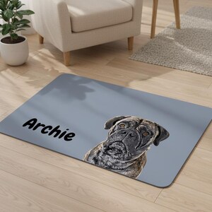 May include: A rectangular, light gray doormat featuring a detailed portrait of a dog with a brindle coat. The dog's name, "Archie," is printed in black cursive on the mat. The mat is placed on a light wood floor.