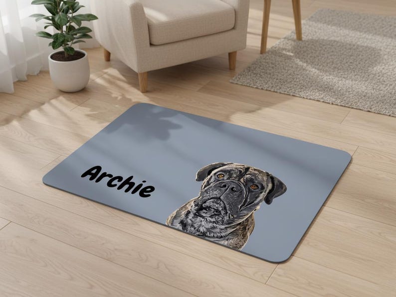 May include: A rectangular, light gray doormat featuring a detailed portrait of a dog with a brindle coat. The dog's name, "Archie," is printed in black cursive on the mat. The mat is placed on a light wood floor.