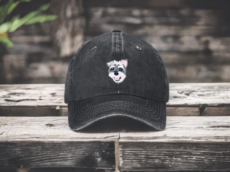 May include: A black baseball cap with a distressed, washed-out finish. The front panel features a small, detailed embroidered graphic of a Schnauzer dog face with grey, white, and black fur, a black nose, and a pink tongue visible in its open mouth.