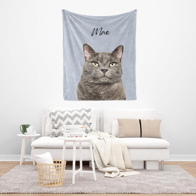 May include: A light blue fleece blanket with a gray cat image and the name "Mae" printed on it. The blanket is displayed on a white couch with pillows and a white side table.
