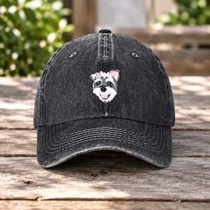 May include: A charcoal black baseball cap with a distressed, washed-out finish. Centered on the front panel is an embroidered patch featuring the detailed head of a Schnauzer dog with gray and white fur, dark eyes, and a pink tongue showing.