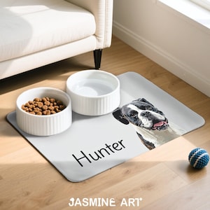 May include: A dog-themed pet feeding station featuring a light gray placemat with a dog portrait and the name "Hunter". Includes two white ceramic bowls, one filled with kibble, and a blue and white ball.