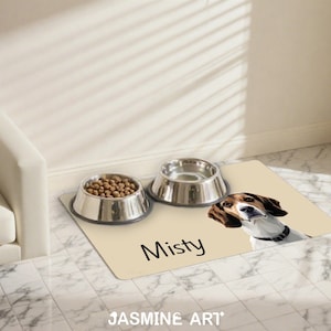 May include: A beige pet feeding mat with a beagle illustration and the name "Misty" in black. Two stainless steel bowls sit on the mat, one filled with kibble. The mat is on a tiled floor.