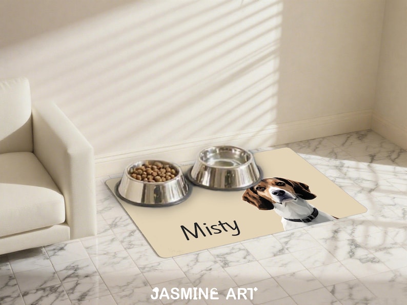 May include: A beige pet feeding mat with a beagle illustration and the name "Misty" in black. Two stainless steel bowls sit on the mat, one filled with kibble. The mat is on a tiled floor.