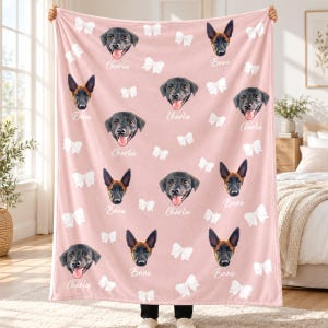 May include: A soft pink fleece blanket featuring a repeating pattern of white bows and portraits of two dogs. The dog faces are labelled with the names Charlie and Bane in white script text, arranged across the fabric surface.