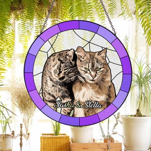 May include: A circular stained-glass window with a purple border, depicting two cats with the names "Sasha & Stella". The cats are in shades of brown and grey. It is designed to be hung, with a chain at the top.