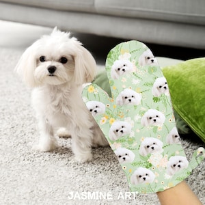 May include: A green oven mitt with a white floral pattern and a repeated image of a white Maltese dog. The mitt has a hanging loop.