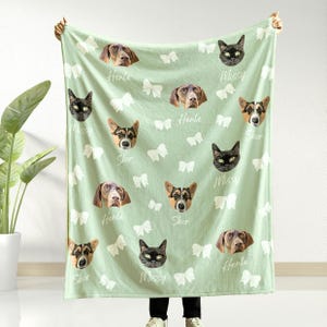 May include: A pale green blanket adorned with dog and cat portraits, interspersed with white bow motifs. The names "Henle", "Missy", and "Skor" are printed near the pet faces. The blanket is held up by a person.