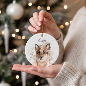 May include: A round, white ceramic Christmas ornament featuring a watercolor portrait of a dog with brown and white fur. The dog's name, "Louie," is written above the portrait in black script. The ornament is held by a person.