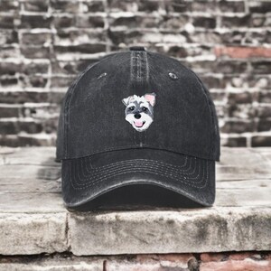 May include: A black baseball cap with a distressed finish featuring an embroidered graphic of a Schnauzer dog face on the front panel. The dog illustration shows a grey and white muzzle, black nose, and pink tongue, centered on the crown.