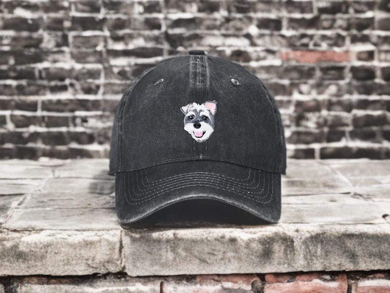 May include: A black baseball cap with a distressed finish featuring an embroidered graphic of a Schnauzer dog face on the front panel. The dog illustration shows a grey and white muzzle, black nose, and pink tongue, centered on the crown.