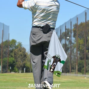May include: A golf towel with a dog graphic, green golf flag, and golf clubs. The towel is light gray with a cartoon dog wearing a golf hat. The word "Loki" is on the hat. The towel is hanging from a person's pants.