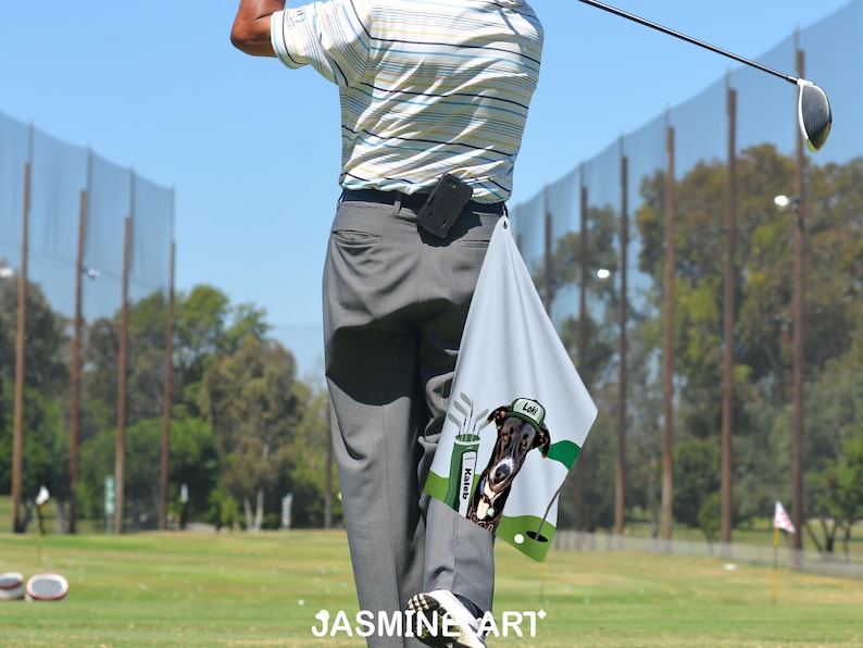 May include: A golf towel with a dog graphic, green golf flag, and golf clubs. The towel is light gray with a cartoon dog wearing a golf hat. The word "Loki" is on the hat. The towel is hanging from a person's pants.