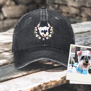 May include: Black denim baseball cap with an embroidered image of a small dog with pink flowers. The dog is wearing a blue collar with the letter "D" on it. The cap has a black brim and a black strap.