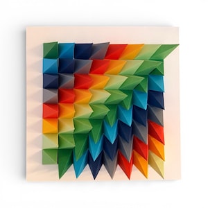 Puede incluir: Una colorida obra de arte geométrico. Presenta una disposición cuadrada de pirámides de papel plegado en un degradado de colores, incluyendo verde, azul, rojo y amarillo. La pieza está montada sobre un fondo blanco.