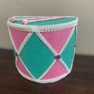 May include: A cylindrical drum covered in pink and teal glitter, featuring a white braided trim arranged in a diamond pattern. Small pink circular gems are placed at the intersections of the trim. Two pink glittery drumsticks rest on top.