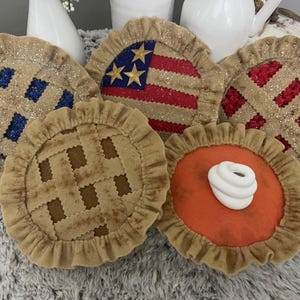 May include: A collection of decorative pie-shaped items. One features the American flag, while others have lattice crusts and various fillings, including blue, red, and orange. Each pie has a ruffled crust and glitter accents, creating a festive appearance.