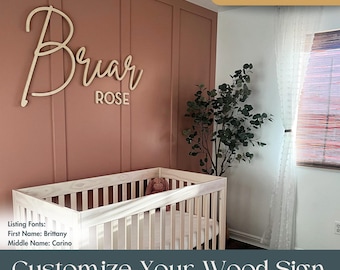 May include: A light-colored wooden wall sign featuring the name Briar Rose in a combination of cursive and block lettering. The sign is mounted on a dusty rose-colored paneled wall above a light wood crib, with a green plant positioned nearby.