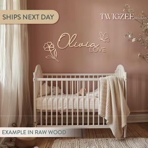 May include: A nursery scene features a white crib with a beige blanket and a wall decoration. The wall decor includes the name "Olivia", a flower, a butterfly, and the word "LOVE", all in a light wood finish. The text "TWIGZEE DESIGNS" is also visible.