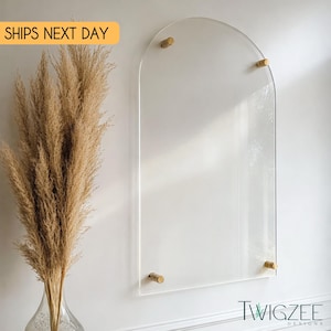 May include: Clear acrylic arch-shaped wall decor with brass-colored hardware. The wall art is mounted on a white wall. A vase of dried pampas grass is to the left of the wall art. The text "TWIGZEE DESIGNS" is at the bottom right.