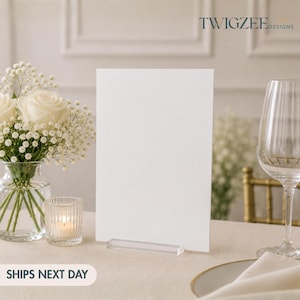 May include: A blank white rectangular sign held by a clear acrylic stand. To the left, a glass vase contains white roses and baby's breath flowers next to a small ribbed glass candle holder. A clear wine glass sits to the right of the sign.