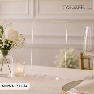 May include: A clear acrylic sign with a rectangular shape and a small base, displayed on a white tablecloth. The sign is next to a vase of white flowers, a lit candle, and a wine glass. The text "TWIGZEE DESIGNS" is visible.