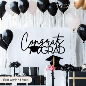 May include: A graduation party scene with black, white, and gold balloons. A large sign reads "Congrats Grad" in black script. Gift boxes are black and white, some with gold accents. A graduation cap is on display.