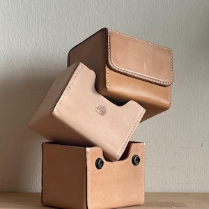 Puede incluir: Tres cajas de cuero apiladas en diferentes tonos de beige. La caja superior tiene un cierre de solapa, mientras que la caja del medio presenta una abertura curva. La caja inferior tiene dos remaches negros. Todas las cajas están cosidas con hilo blanco.