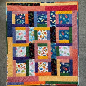 May include: A colorful patchwork quilt featuring various sea creature patterns. The quilt showcases squares with jellyfish, fish, and other marine life in vibrant hues of blue, green, orange, and purple. The quilt's dimensions are approximately 60 inches by 70 inches.
